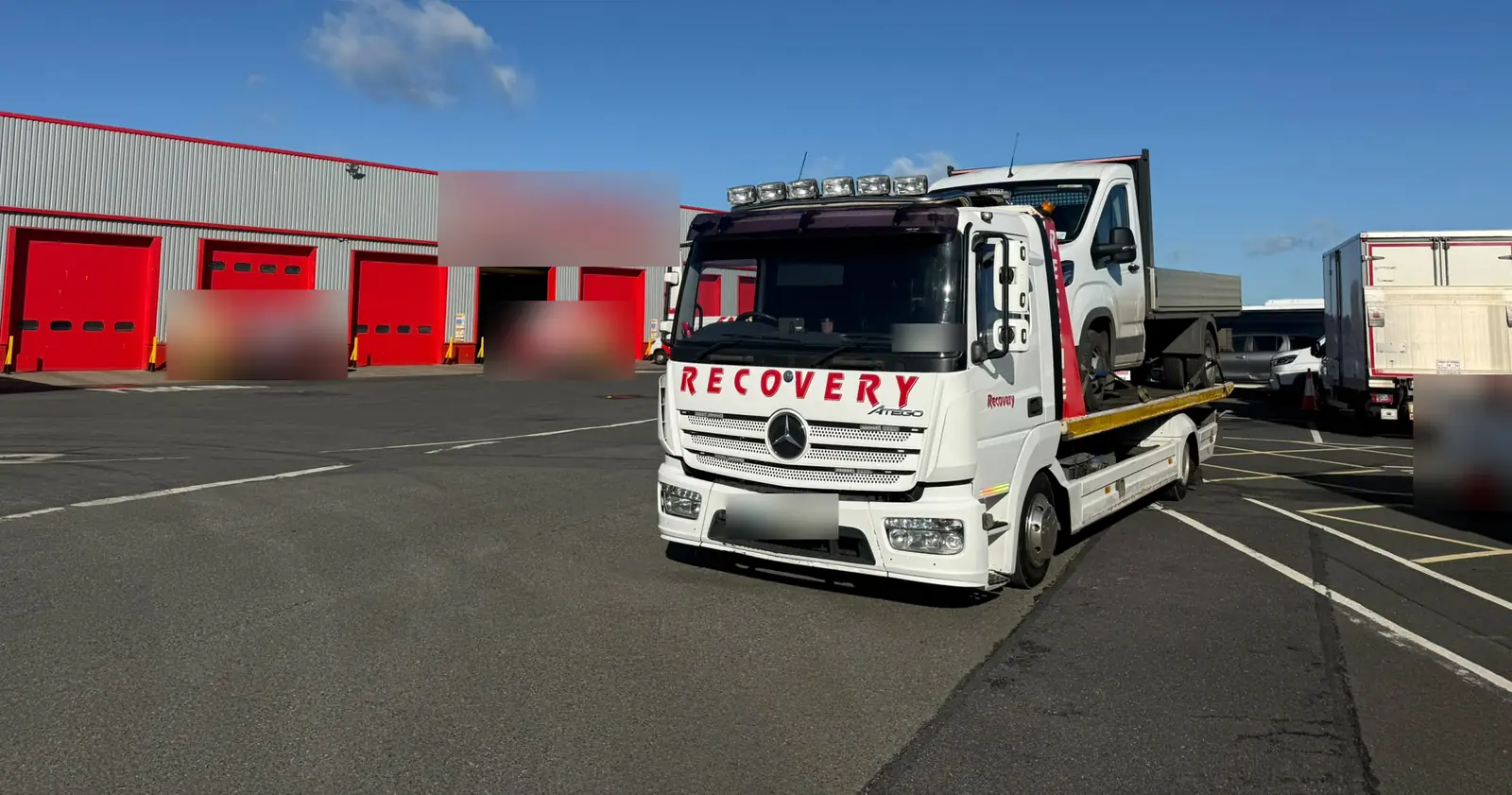 Front view of a Recovery CC Mercedes Atego tow truck carrying a dropside pickup in a sunny yard