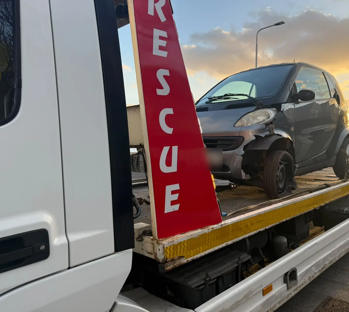 A small city car with front damage loaded behind the red RESCUE post of a Recovery CC tow truck at sunset