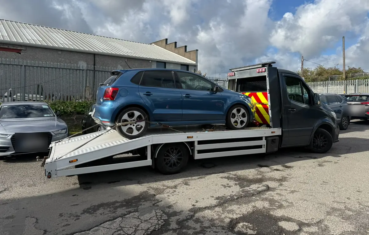A blue hatchback on a dark flatbed recovery truck in a Dublin yard