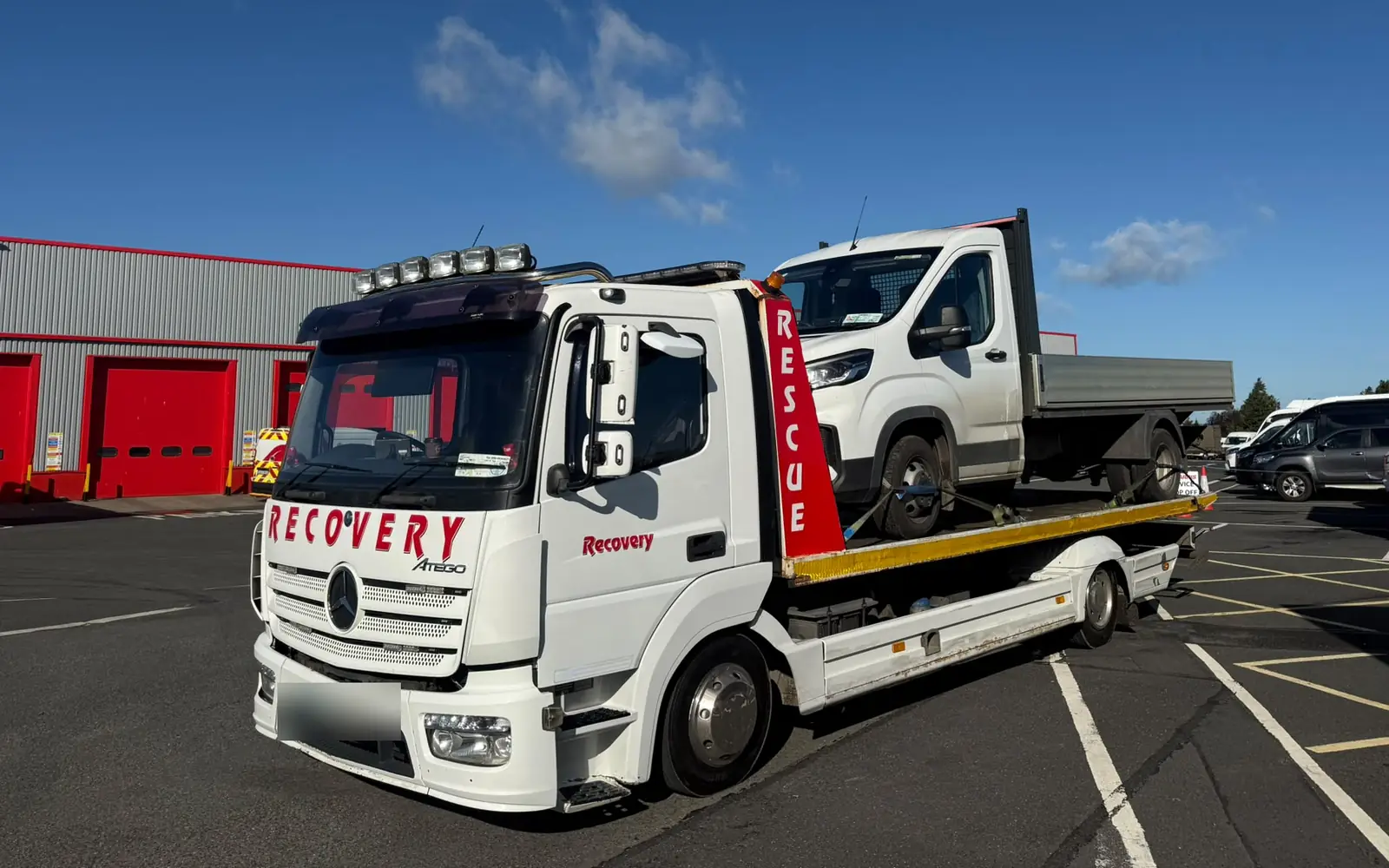 A white Recovery CC Mercedes Atego recovery truck carrying a van, parked in the sun