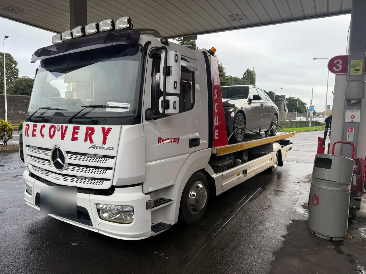 A white Recovery CC truck with a car on the deck, parked on a wet forecourt