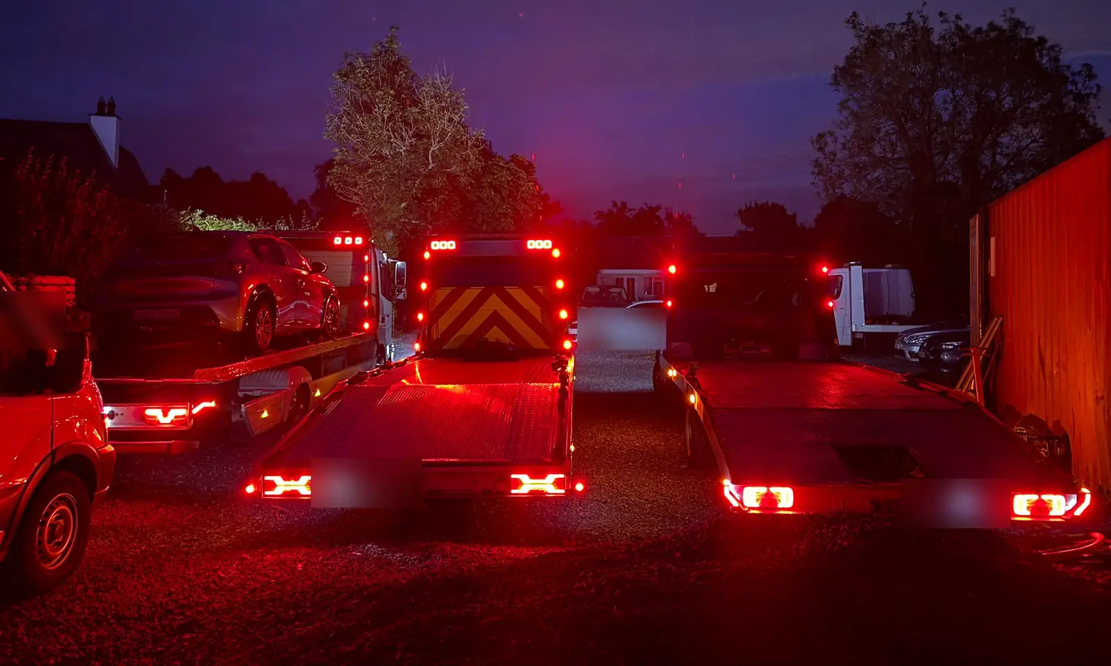 Two Recovery CC tow trucks seen from behind at dusk, rear lights glowing red