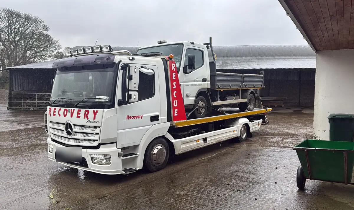 A Recovery CC Atego tow truck with a small tipper truck loaded on the deck in a yard