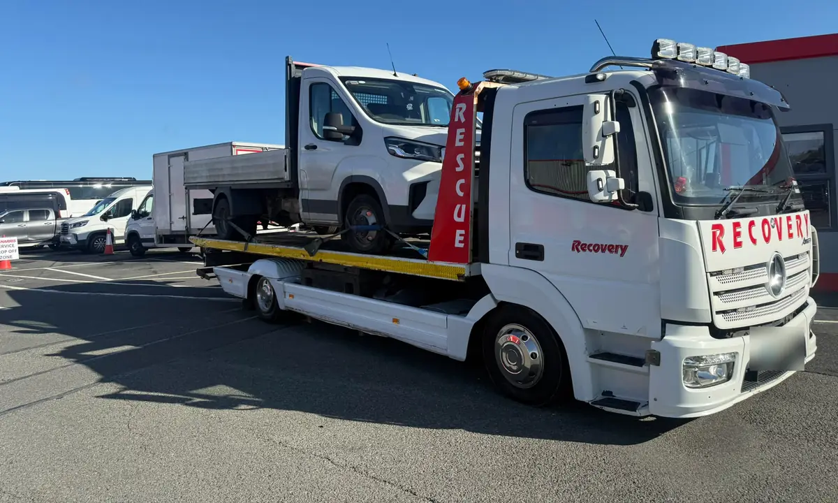 A Recovery CC tow truck carrying a white tipper van across a sunny car park